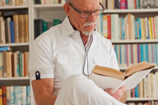 Egy idősebb férfi könyvespolc előtt könyvet olvas.