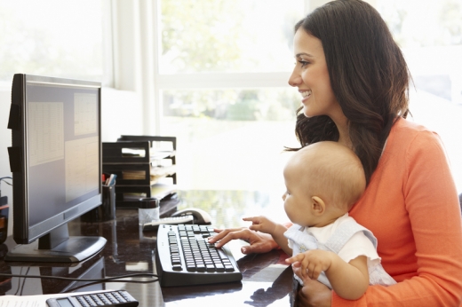Egy nő a számítógép előtt ülve, egyik kezével gépel, másikban a kisbabáját tartja.