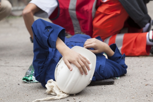 Egy munkás sérülten fekszik a földön és a mentős fölé hajolva segít neki.