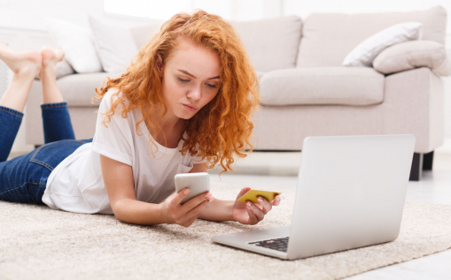 Fiatal vörös és göndör hajú lány a padlón fekszik és laptopot használ. A kezében van egy bankkártya és egy telefon.